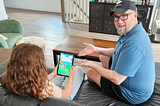 Father sitting beside daughter who is playing a mobile game on a tablet