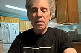 An older man with gray hair wearing a black t-shirt that has partially visible text reading “DON’T GET CAUGHT.” He is seated in a well-lit kitchen with wooden cabinets and a refrigerator in the background. The refrigerator has decorative magnets, including butterfly designs. The man appears to be looking downward with a neutral or contemplative expression.