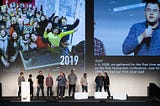 IxDA Regional Coordinators on stage in Milan, Italy at Interaction 20. Behind them are images on screen from the Local Leaders retreat and a close up of Jack Moffett with the microphone.