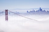 Photo of fog overflowing the Golden Gate Bridge and invading the city. You can see one tower of the bridge in muted red, with cables sweeping down and away from it, artistically, and disappearing into the fluffy white. In the background is the bluish charcoal grey skyline of San Francisco. The sky is a dull blue getting ready for twilight. You might think of the fog as a blanket, but it’s chilly rather than cozy.