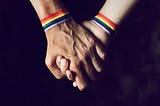 Two white men wearing rainbow wristbands holding hands over a black background