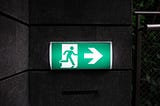 Photo of a green exit sign on grey brick wall. Sign has an icon of a person going through a door and a white arrow pointing to the right.