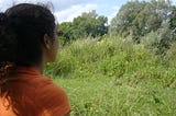 Woman in orange t-shirt looking at lush grassy field and blue sky.
