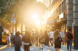 Many people walking on a city street, with sunlight bursting through a tree