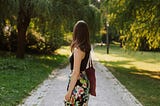 A woman walking alone on a narrow path surrounded by trees, with her back to the camera.