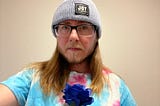 The author is wearing a knitted blue hat reading “Joy as Resistance”, black rimmed glasses, and a blue, white, and pink shirt. He is holding a dark blue flower. He has a beard and mustache and a stern look with a crease between his eyebrows.