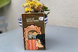 Paperback copy of Love of Seven Dolls by Paul Gallico resting on a flower pot on an outdoors table.