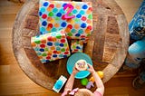 photo shows a table with a pile of presents wrapped in balloon wrapping paper with the number 2 on top. In front of the gifts, a pair of small arms reach over some discarded toys to hold a vanilla cupcake with sprinkles