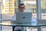 A man in his mid 20s wearing a white sweather and blue jeans. He’s sitting on a chair. He’s resting a MacBook against a glass table. He has black hair and glasses. Behind him is a window and several skyscrapers.