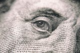 A photo by Vladislav Reshetnyak from Pexels zooming in on Benjamin Franklin’s eye from a one hundred dollar bill.