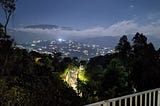 From balcony of house in a mountain- a road bathe in street light then further city light below clouds on distant hill