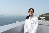 Jane Wong, dressed in a white coat, stands on a balcony at the HKUMed campus overlooking the sea