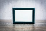 An empty picture frame by a white wall, standing on a wooden top.
