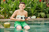 Image of a preteen playing the ukulele with a coconut drink beside him with his legs dangling in the pool