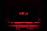 An image of the Netflix logo on a television screen in a dark room. The television is on top of a cabinet with a console gaming controller sitting in front of it. The red from the logo font resonates outward from the television giving a sort of ominous glow.