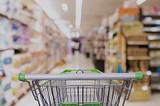 An image of a grocery cart standing in the middle of a grocery store aisle, from the perspective of someone pushing the grocery cart. The cart is in focus and the products on the shelves are blurred out.