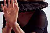 Dancer Molly Heller poses for the camera, hands gestures close to the camera. She wears a black shirt and a tutu-like horizontal folded collar around her neck.