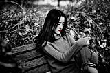 A black and white image of a sad Asian woman on a park bench. Her lips are red.