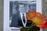 Black and white photo of a memorial brochure, showing Alex,  who died by suicide in 2012. With a yellow rose from the author’s garden…