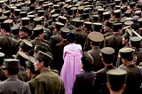 A woman stands in a crowd of North Korean soldiers wearing a pink Hanbok