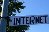 A plate in white at a mid length public light post which is written, in black, the word internet; a blue sky behind; tree top