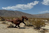Rust colored herbivore dinosaur sculpture in the desert with gray mountains in background and blue sky with a few white clouds.