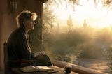 A writer sits quietly on a desert patio at sunrise, looking toward the Sonoran Desert while an open notebook and untouched pen rest beside a cup of coffee, suggesting reflection before writing.