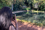 A girl taking a picture of an artificial pond with overgrown creepers dipping in.
