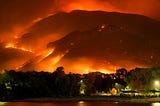 Rings of blazing fire engulf mountains  surrounding a town and reflect in the adjacent lake.