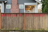 The words “We are all in the this together” are painted in large red letters on a brown fence in front of a grey house.