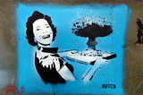 Photo of street art on a concrete wall. Against a blue background, a smiling 1950s housewife in black and white carries a serving tray containing a mushroom cloud. Via Wikimedia Commons/James Nash (aka Cirrus), license CC 2.0.
