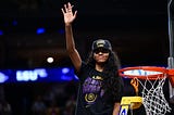Angel Reese waving to the crowd after winning the NCAA Title. She is wearing a Black “National Champs” hat and shirt and is standing on a yellow ladder next to the basketball rim.