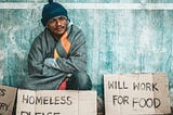 Homeless man with back against a wall and supplication signs — one says “WILL WORK FOR FOOD”.