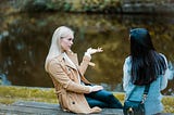 Two women sitting on a bench beside water, talking.