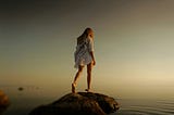 A woman stands barefoot on a rock by calm waters at sunset.