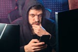 Man with a mustache in a dark hoody, holding a smartphone and looking dubious