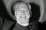 Uma foto preto e branco de Robert Anton Wilson, um homem branco, de origem irlandesa, um pouco careca, com um pouco de barba, pelos brancos, sorriso no rosto e roupas escuras.