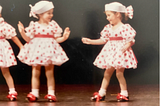 A young Riley Young stands on stage wearing a white dress with red polka dots, two other fellow dancers stand beside her