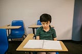 A young boy sits alone at a school desk, looking down at an open but empty notebook. He wears a dark green polo shirt and appears thoughtful or sad. Bright blue chairs and beige desks surround him in a plain classroom setting.