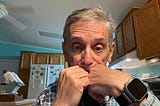 An older man sits in a kitchen, resting his face against both fists in a thoughtful or concerned pose. He is wearing a plaid shirt and an Apple Watch. The background includes wooden cabinets, a white refrigerator with butterfly magnets, and teal-colored walls.