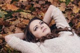 Young Attractive Brunette Woman In White Jumper Lying Down On Grass With Autumn Leaves.