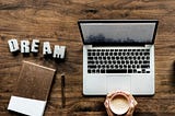 A person is holding a cup of coffee with both hands infront of a laptop. The word dream is pinned on the desk with a diary