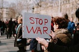 A person, whose face is out of frame, holds a signs that reads “STOP WAR” during a gathering of people outdoors. There are old neo-classical buildings and trees without leaves in the background.