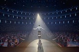 A spotlight shines on a perfomer on a stage in front of a large audience of people. This performer, it turns out, is a monkey.