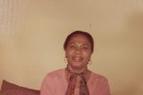 My grandmother sitting on a sofa, wearing a pink shirt and brown print scarf.