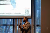 Dr. Jackie Beard in a gray blazer stands at the front of a conference room, holding a microphone and addressing attendees. Behind him, a large projection screen displays a presentation for the Association of Florida Colleges Region IV Conference, while floor-to-ceiling windows with vertical blinds line the room