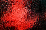 A window filled with droplets of water and red and turquoise light emanating from the other side.