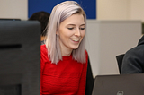 Photo of Natasha smiling and wearing a red jumper whilst sat at a desk typing on a laptop.