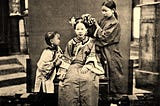 A lady sits still while a girl dresses her hair and a child tries to get her attention.