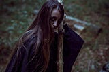 Mysterious Woman / witch with long dark hair and black robes and a shadowy face holds a staff that is partly charred, background of woodland